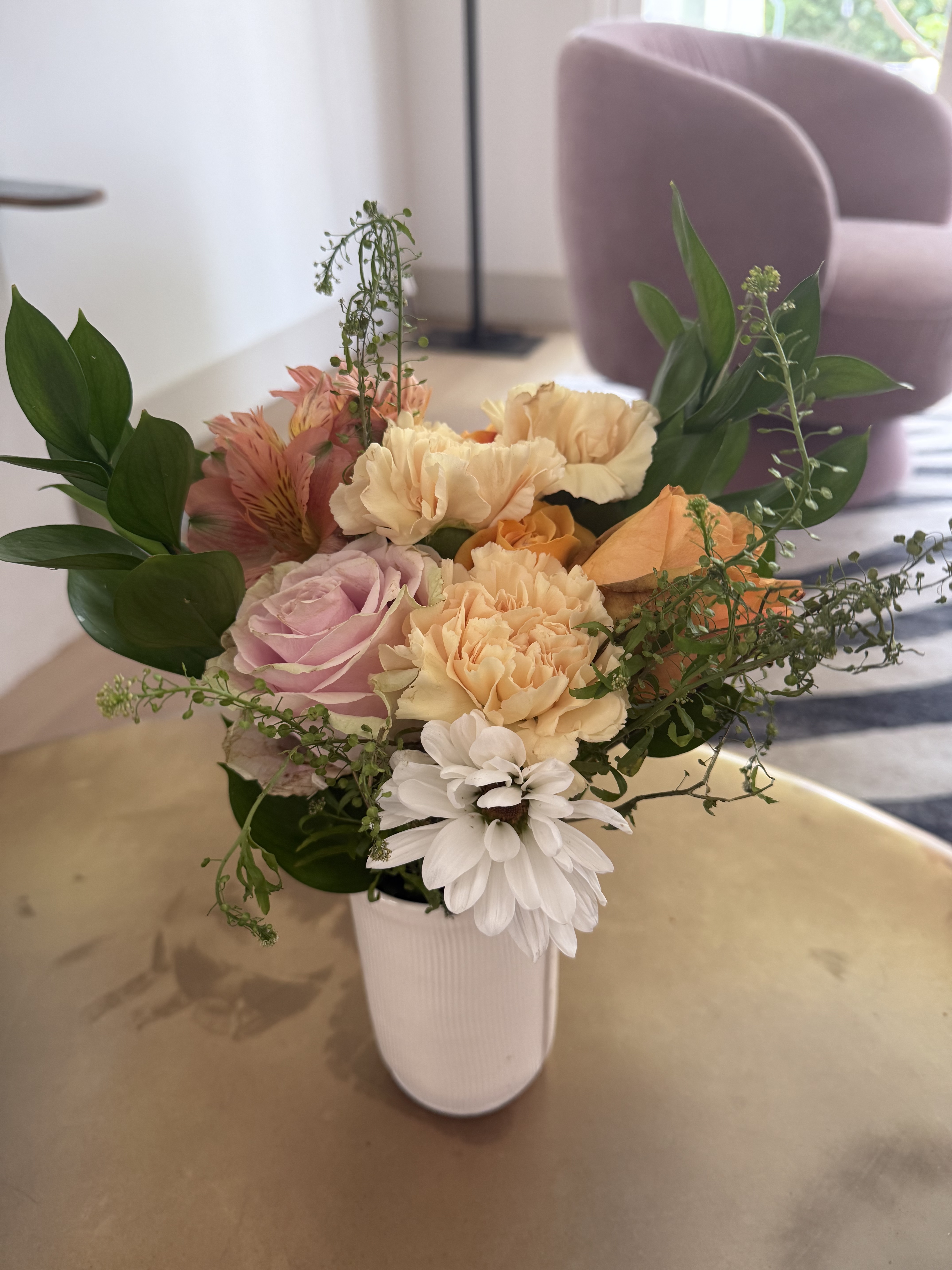A bouquet of peach carnations, lavender roses, alstroemeria and greenery in a white ribbed vase